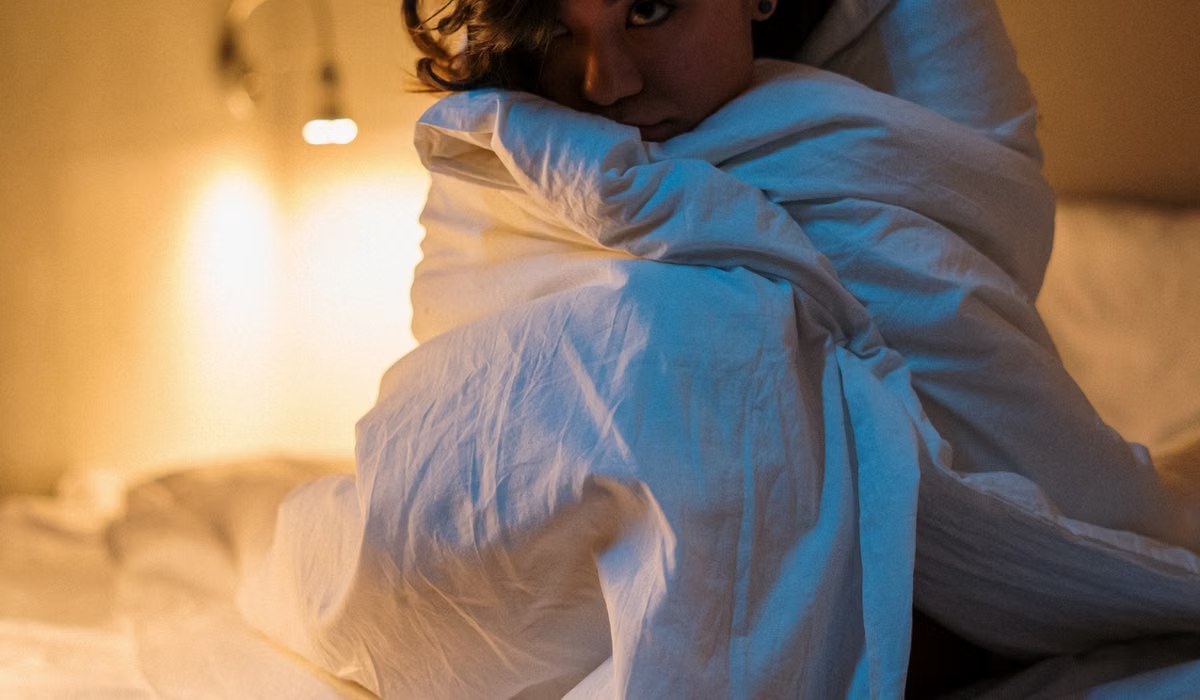 Person wrapped in a white duvet in a bedroom lit by a warm bedside lamp