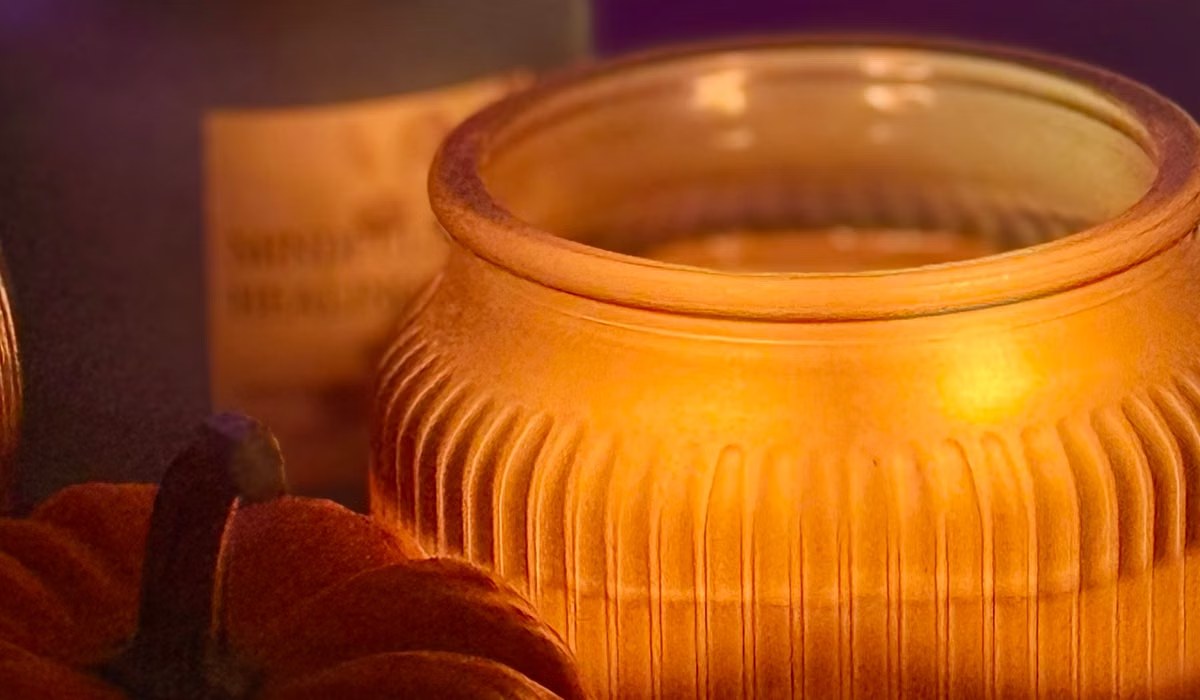 Glowing candle in a ribbed glass jar next to a small pumpkin on a woven tray