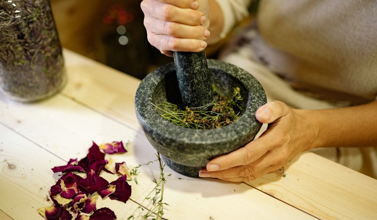 Hands grinding dried herbs and rose petals in a stone mortar and pestle on a wooden table