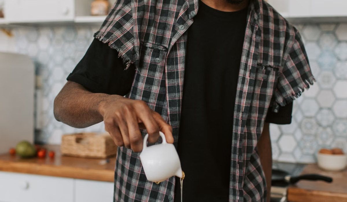 Person in a plaid shirt pouring syrup from a small white pitcher in a bright kitchen