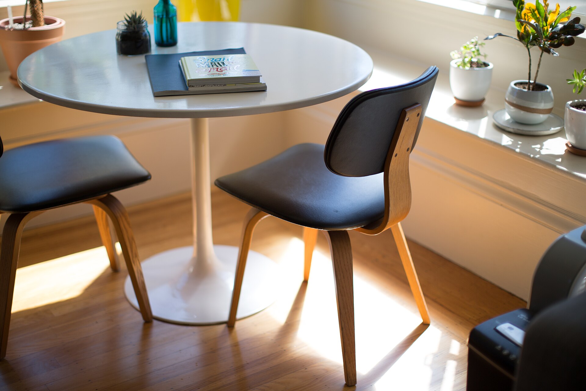 Sunlit breakfast nook with a round white tulip table, wooden chairs and potted plants on the windowsill